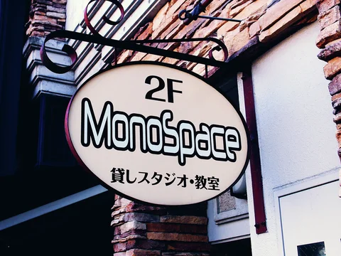 An oval MonoSpace studio sign hangs from decorative black scrollwork beside brick and pale wall panels.