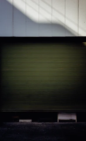 A diagonal shadow crosses pale upper panels above a closed green shutter and a dark pavement edge.