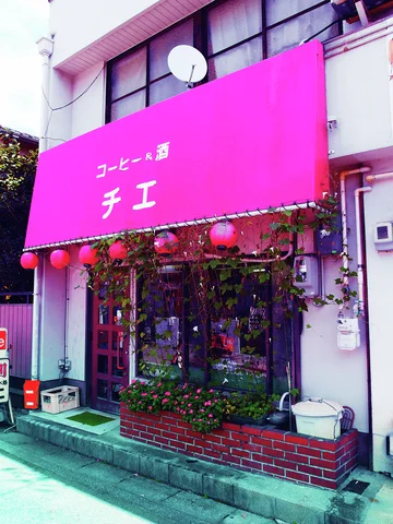 A vivid pink awning shelters a small cafe entrance bordered by climbing plants and red lanterns.