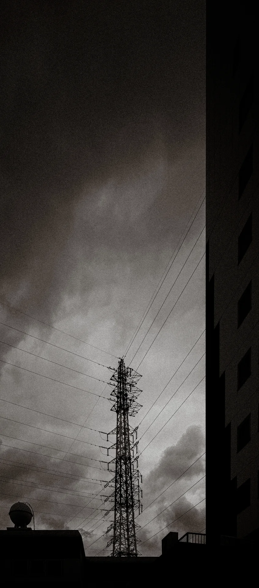 A lattice electricity tower rises beneath cloudy sky beside the dark edge of a tall building.