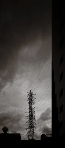 A lattice electricity tower rises beneath cloudy sky beside the dark edge of a tall building.
