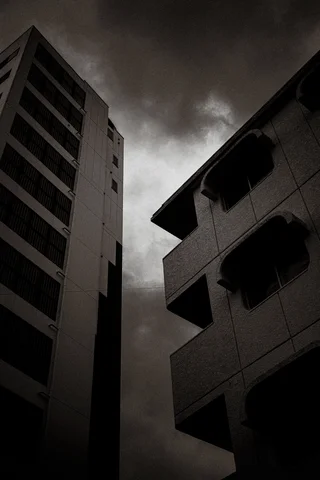 Tall buildings frame a narrow strip of cloudy sky above deep shadow in black and white.