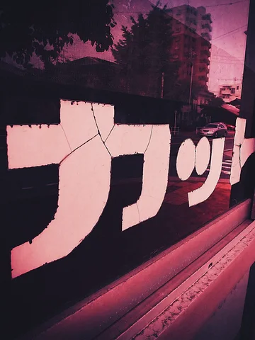 Large pale Japanese lettering crosses a dark reflective window tinted pink by surrounding buildings and foliage.