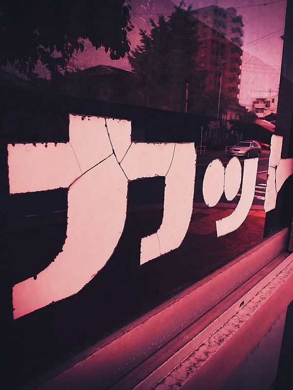 Large pale Japanese lettering crosses a dark reflective window tinted pink by surrounding buildings and foliage.