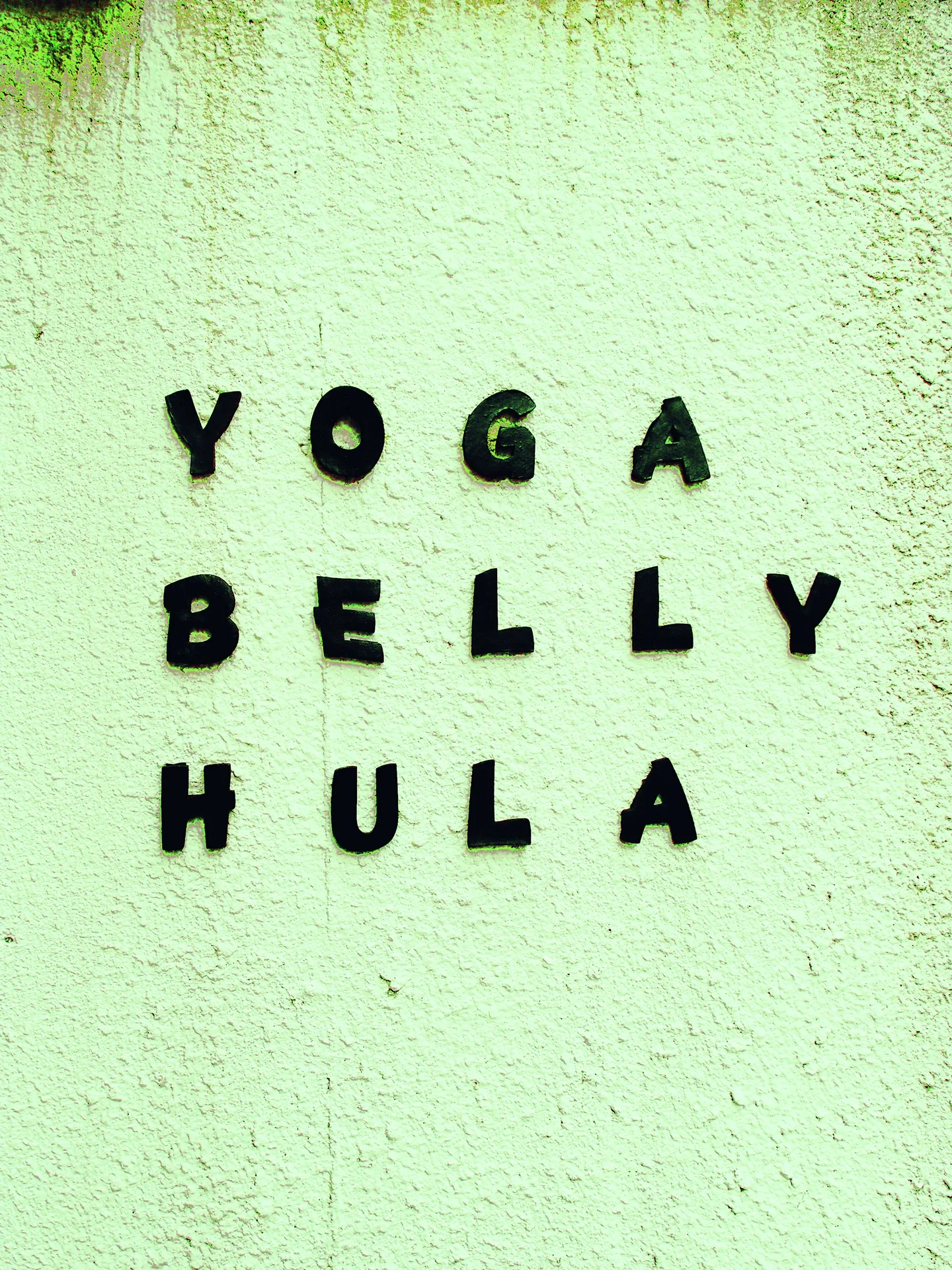 Small black letters spelling Yoga, Belly, and Hula form three rows on a pale green textured wall.