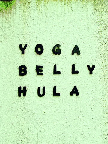 Small black letters spelling Yoga, Belly, and Hula form three rows on a pale green textured wall.
