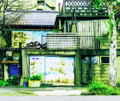 Rust-stained pale doors sit below a cluttered balcony beside a tree and weathered wooden walls.