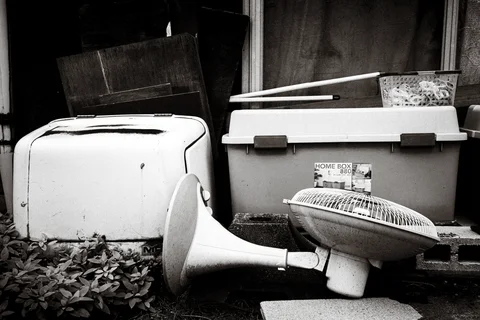 Discarded appliances and a plastic storage box crowd a shaded corner in black and white.