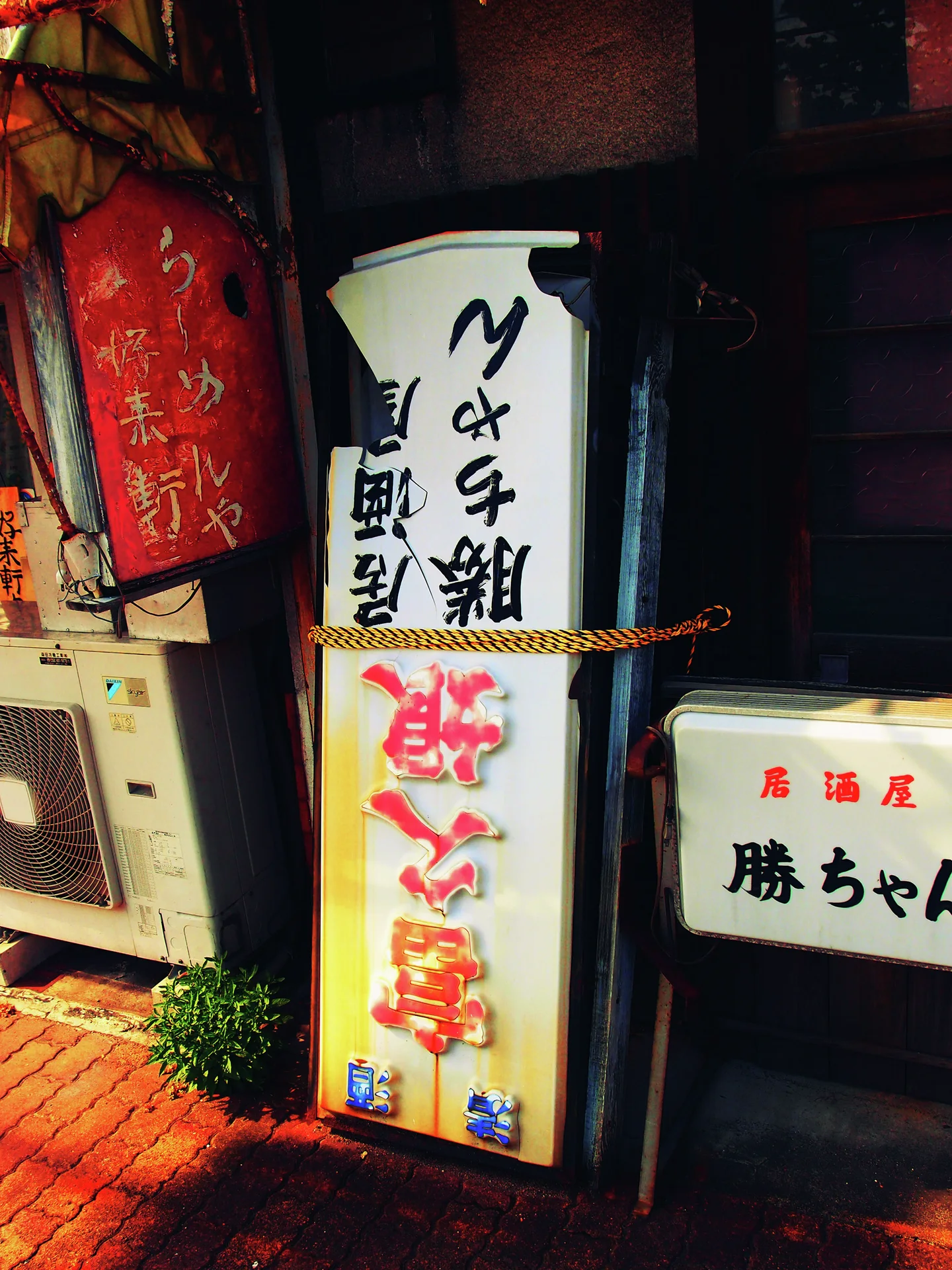 Several illuminated Japanese signs cluster beside a dark storefront entrance and an outdoor air conditioner.