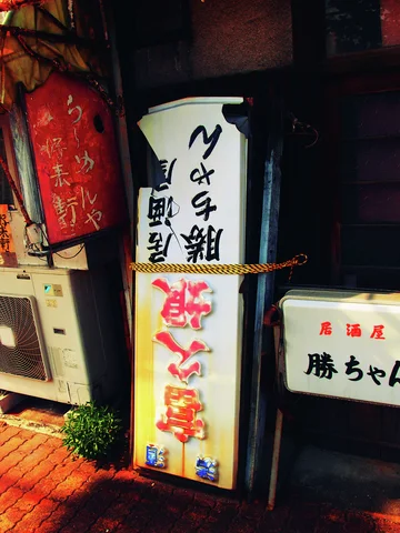Several illuminated Japanese signs cluster beside a dark storefront entrance and an outdoor air conditioner.