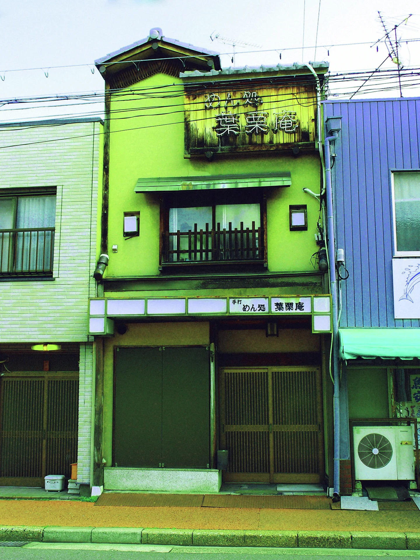 A narrow green storefront has a dark closed entrance beneath a small balcony and weathered upper sign.