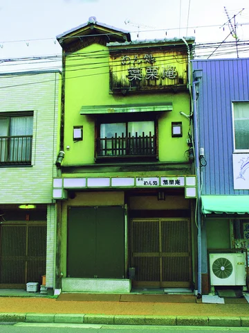 A narrow green storefront has a dark closed entrance beneath a small balcony and weathered upper sign.