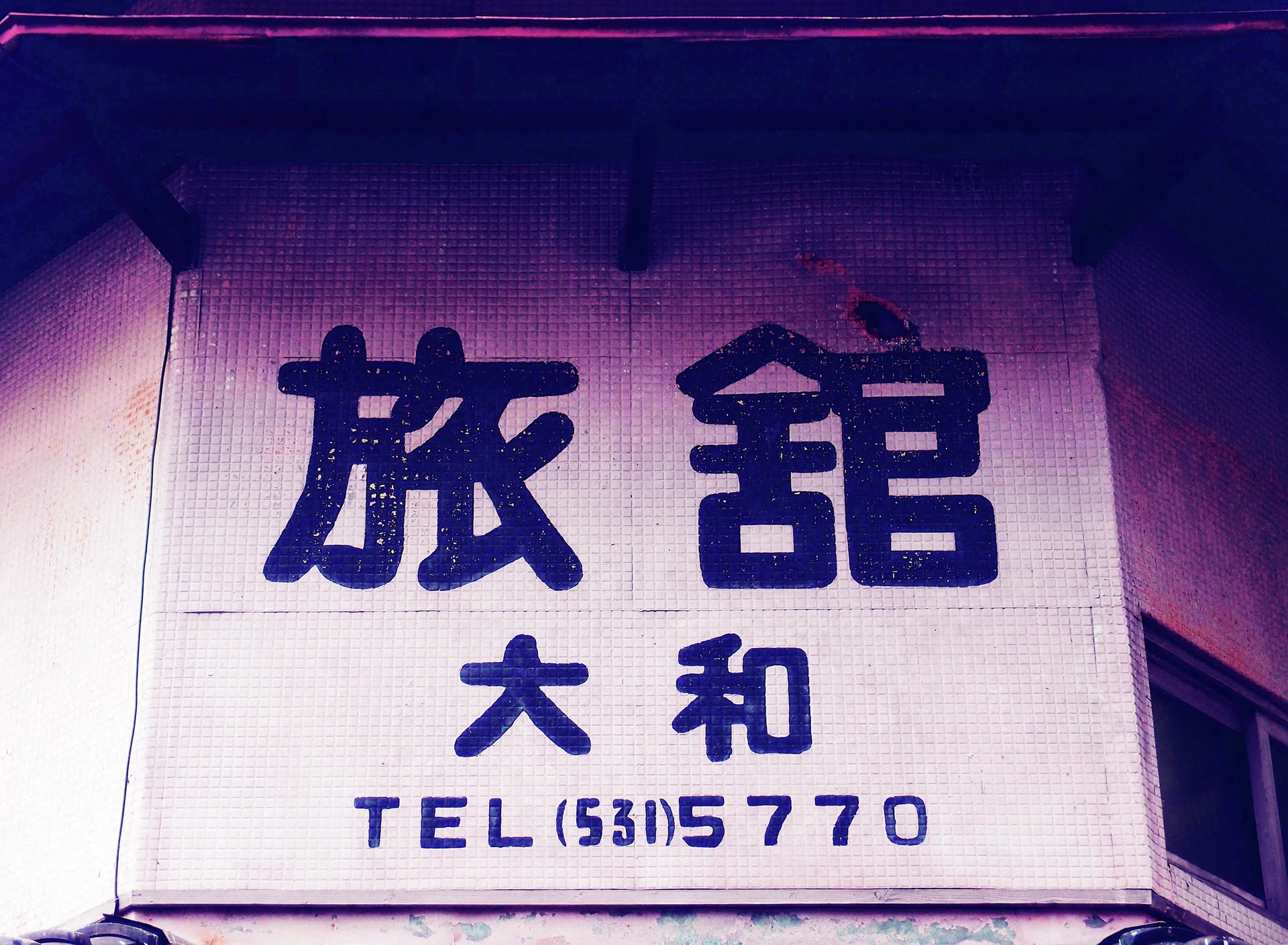 Large dark Japanese characters and a phone number cover a pale tiled wall beneath deep eaves.