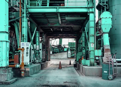 Turquoise industrial columns and pipes surround an open passage with traffic cones and a small handcart.