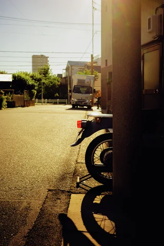 A parked motorcycle casts a curved shadow beside a sunlit open lane and shaded building corner.