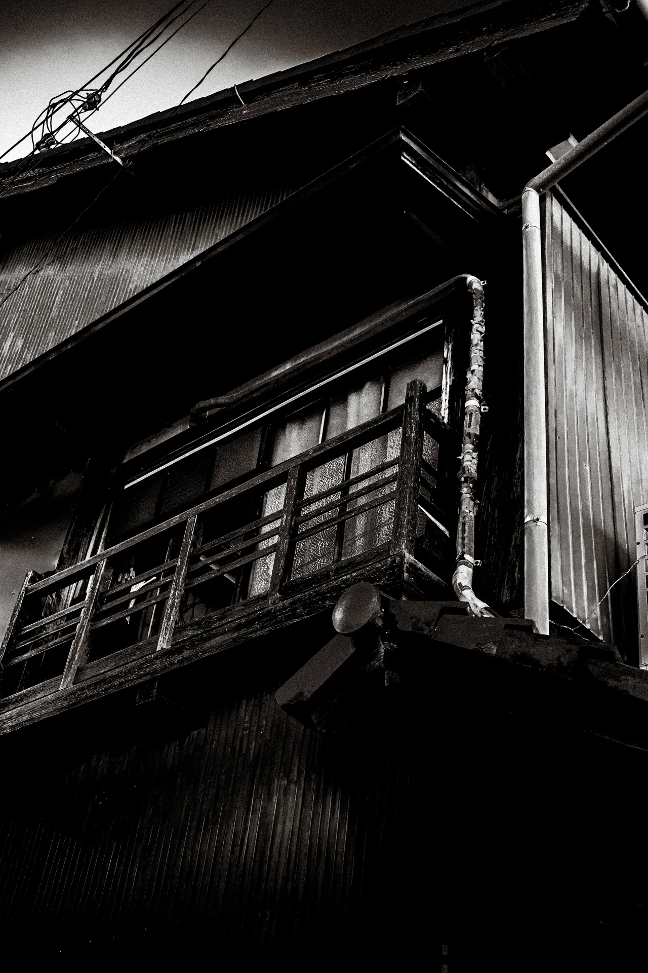 An old wooden balcony with metal railings projects from a shadowed house in monochrome.