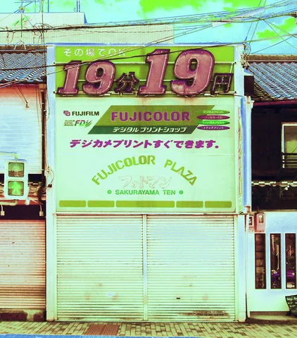 A closed storefront has a pale green shutter beneath a large sign with pink numbers.
