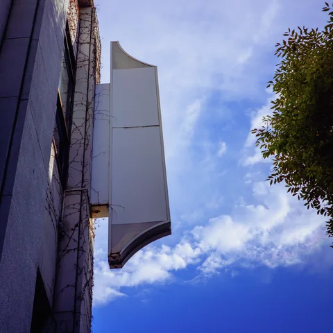 A blank white projecting sign extends from a gray wall beside blue sky and leafy branches.