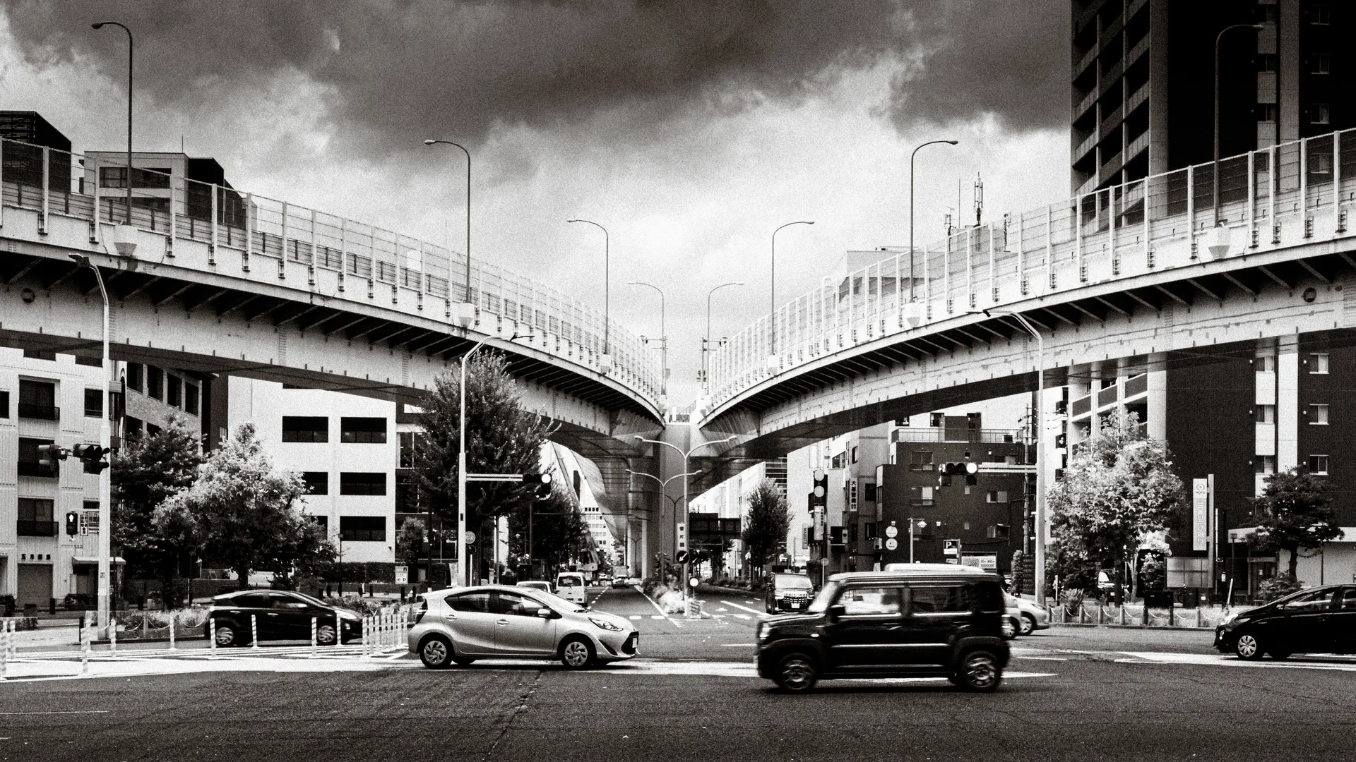 Cars cross beneath two curving elevated roadways under dark clouds in black and white.