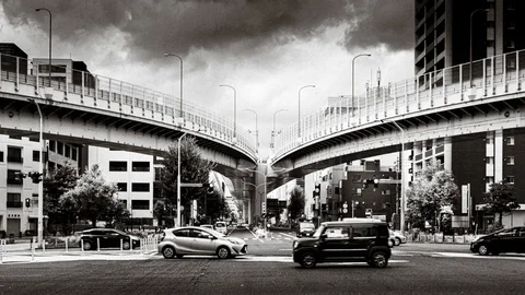 Cars cross beneath two curving elevated roadways under dark clouds in black and white.