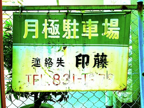 A weathered green and white parking sign hangs on chain-link fencing beside plants.