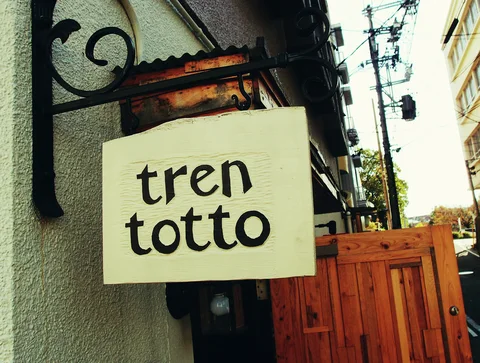 A pale tren totto sign hangs from black scrollwork beside a textured wall and orange wooden gate.