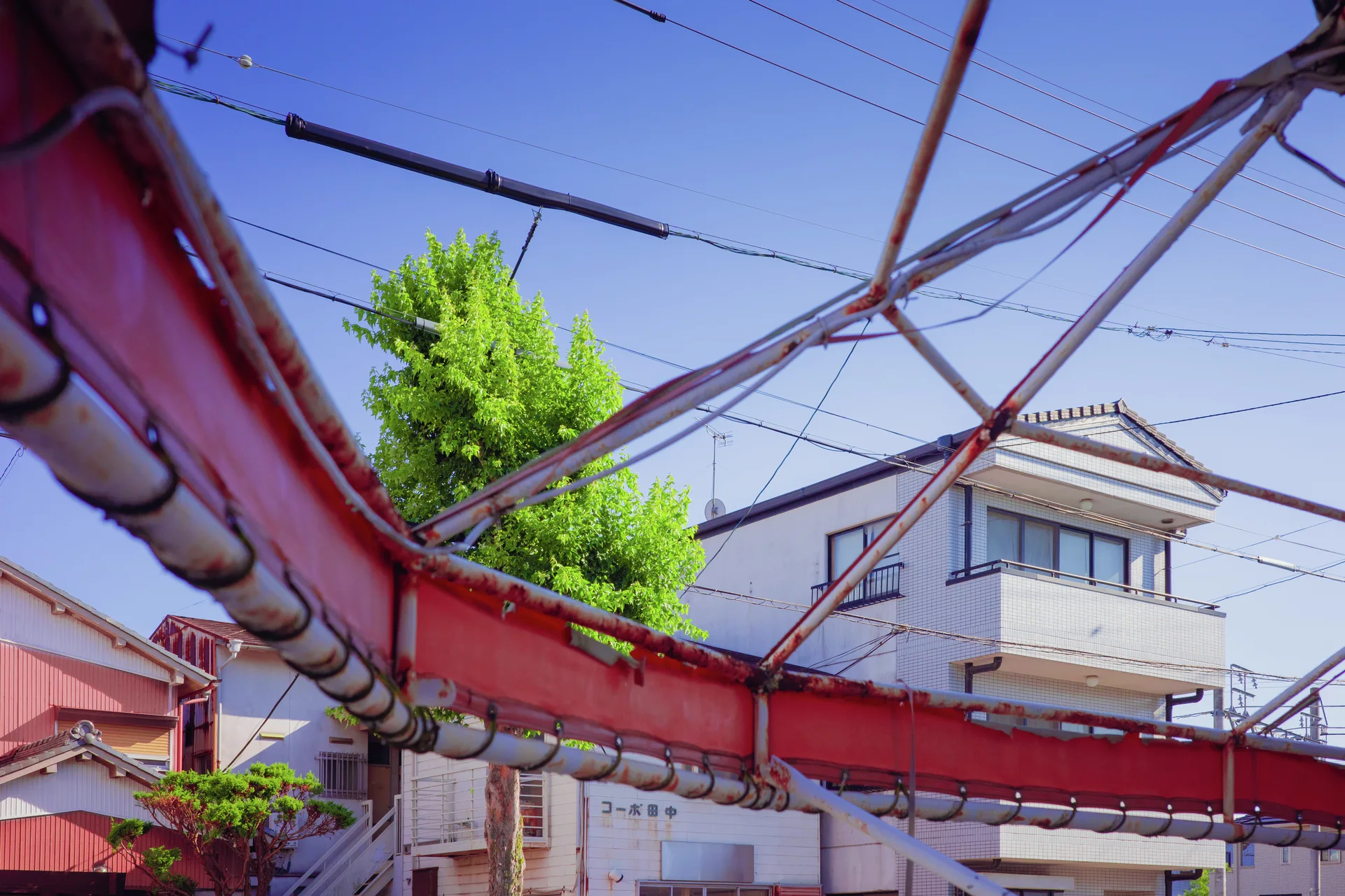 Red metal beams and crossing braces frame a leafy tree and pale house beneath overhead wires.