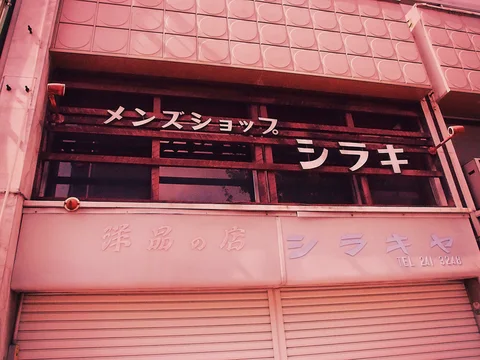 White Japanese lettering spans dark upper windows above closed pink shutters and faded facade signage.