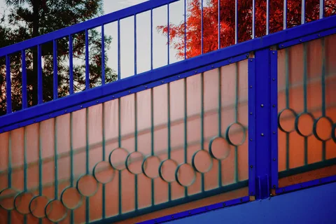 Circular metal motifs cross translucent orange panels within a vivid blue fence beneath red-leaved branches.