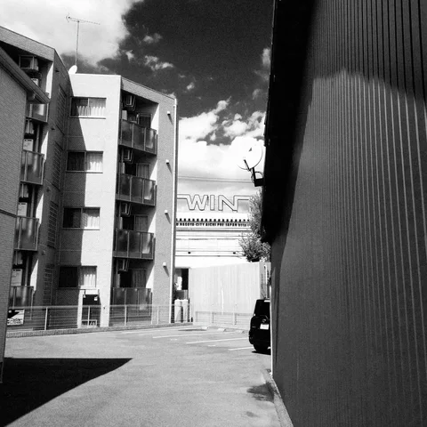 A narrow street passes apartment balconies beside a dark corrugated wall beneath high-contrast clouds.