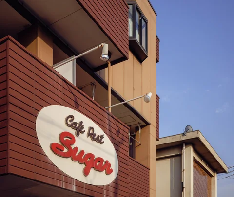 An oval cafe sign with raised red lettering hangs on a brown facade beneath two projecting lamps.