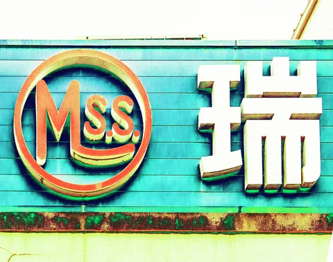 An orange circular emblem and large pale Japanese character project from a turquoise sign above rusted trim.