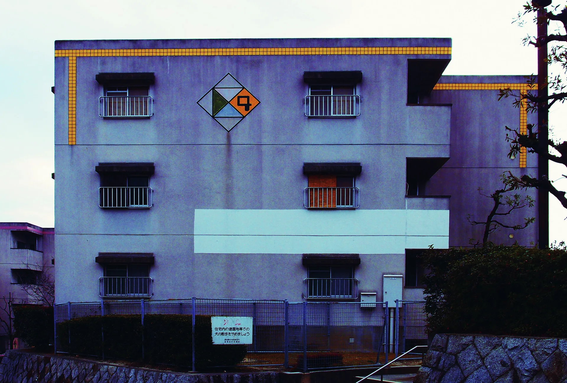 A pale apartment facade has rows of barred windows, yellow roof trim, and a colored diamond emblem.