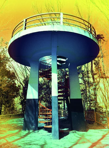 A circular elevated platform with railings rests on blue supports around a spiral staircase among trees.