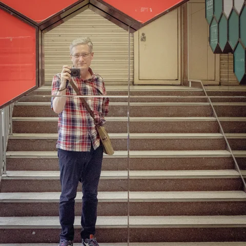A person in a plaid shirt holds a camera before stairs reflected in glass.