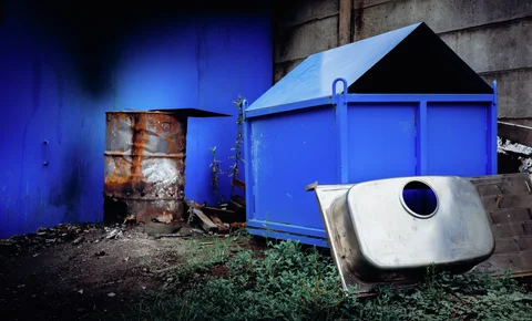 A discarded metal sink leans beside a blue industrial container and rusty barrel.