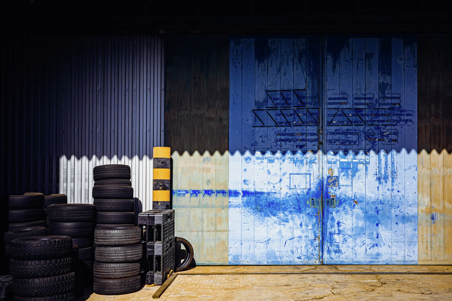 Stacks of tires stand beside a corrugated door covered with weathered blue and white paint.