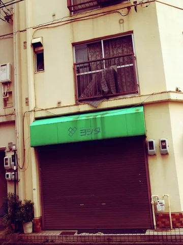 A closed dark shutter sits beneath a faded green awning and large barred upper window.