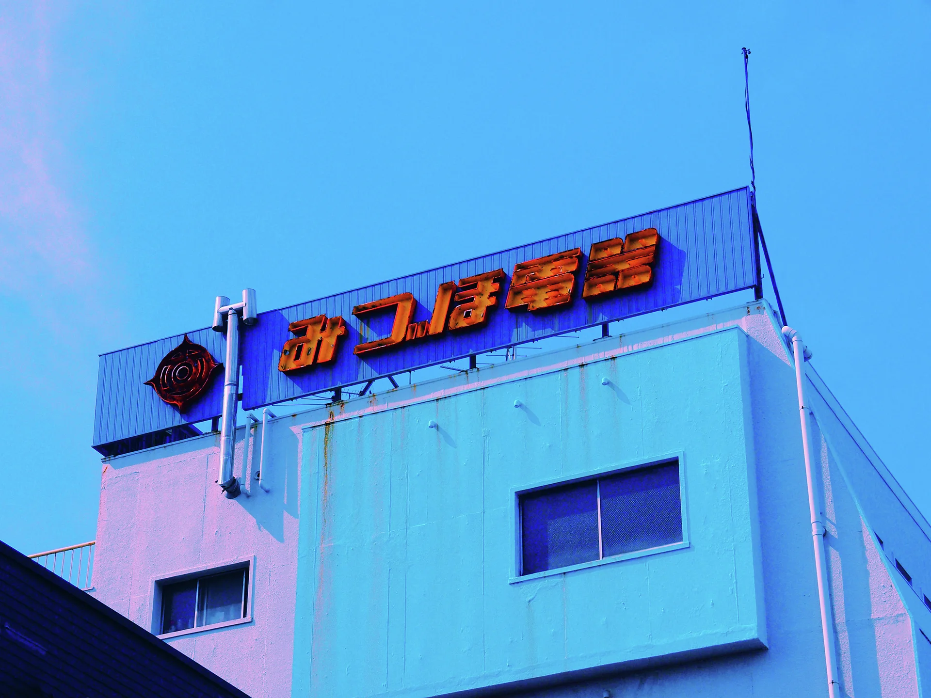 Orange Japanese lettering tops a pale blue building with two recessed windows beneath a vivid blue sky.