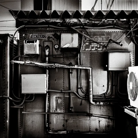 Meters, boxes, and tangled cables cover patched metal siding beneath a corrugated awning in black and white.