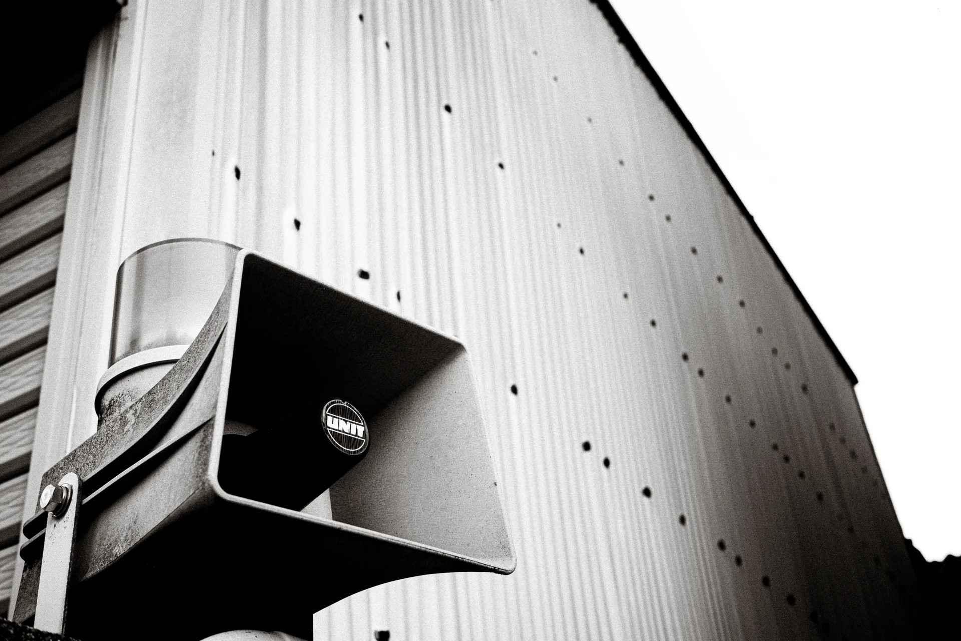 A square horn loudspeaker projects from a corrugated metal wall in an upward black-and-white view.