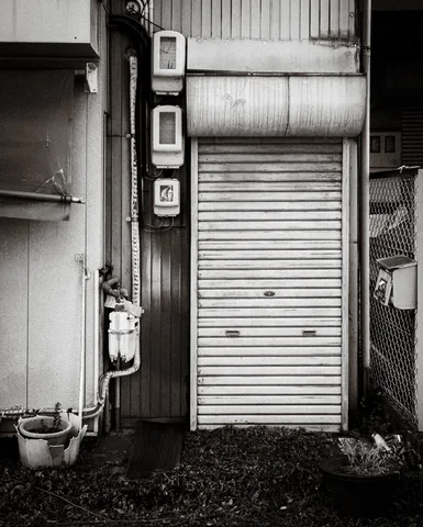 A narrow closed metal shutter stands beside exposed pipes and small wall fixtures in black and white.