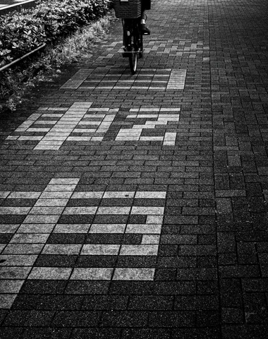 Large pale characters are set into dark brick pavement beneath a distant cyclist in black and white.