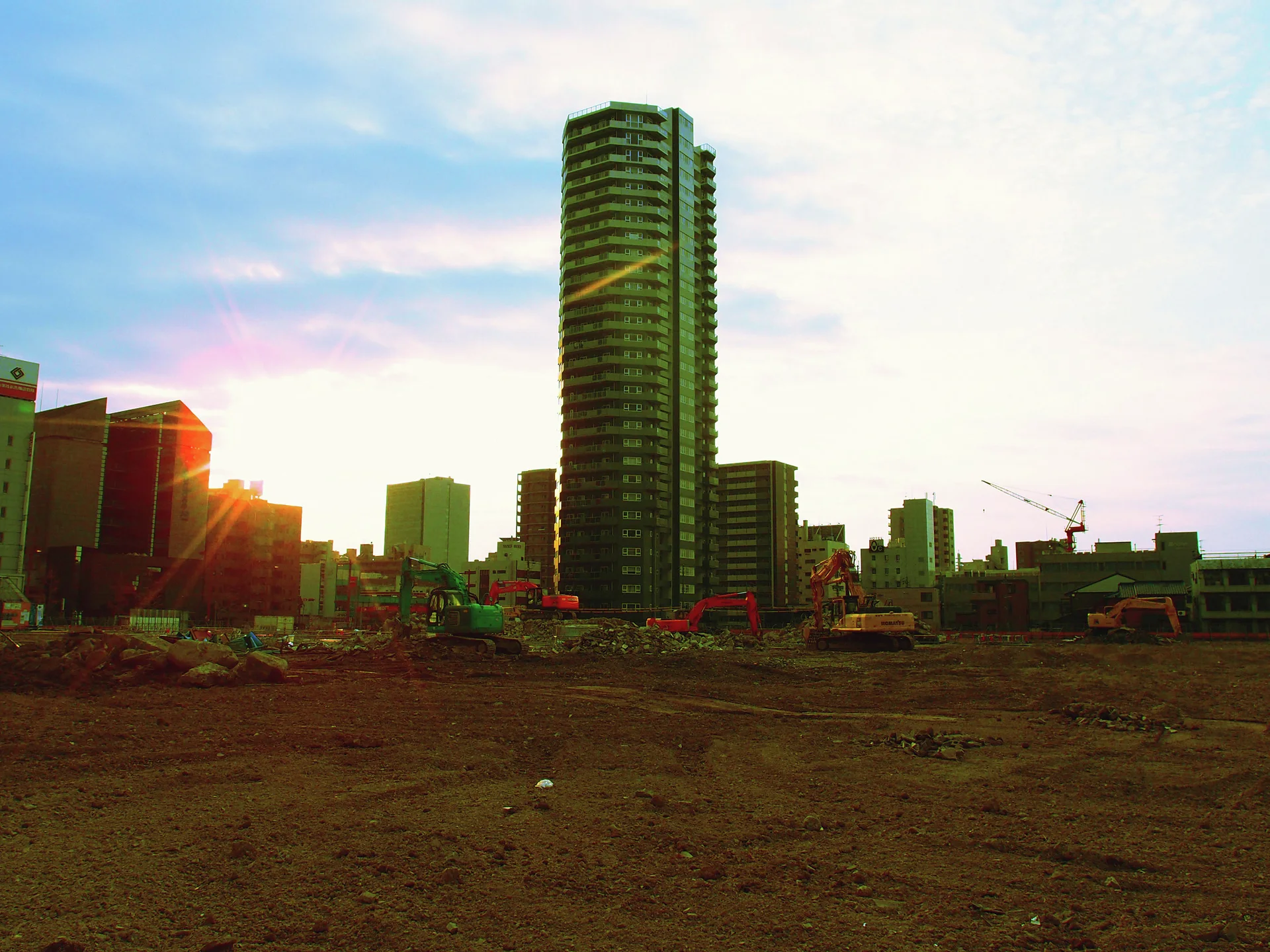 A tall apartment tower rises beyond an open earth-covered site with low sunlight near the horizon.