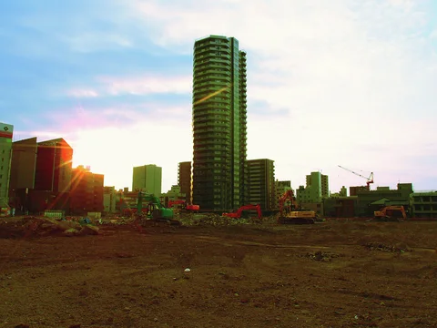 A tall apartment tower rises beyond an open earth-covered site with low sunlight near the horizon.