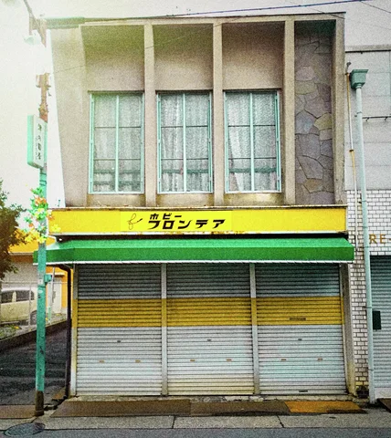 Closed gray shutters with a yellow band sit beneath a green awning and three tall upper windows.