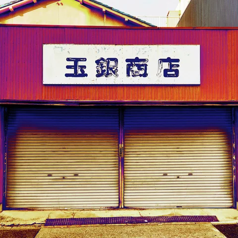A white Japanese sign hangs above two closed cream shutters on a reddish corrugated storefront.