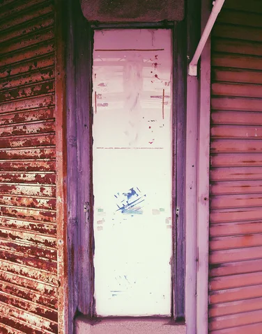 A narrow pale door stands between worn reddish metal shutters with peeling paint and dark trim.