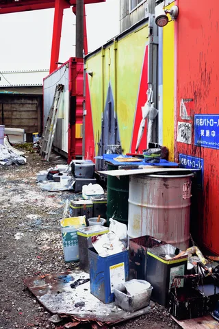 Metal drums and assorted containers line a cluttered industrial passage beside yellow and red walls.