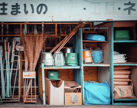 Turquoise shelves hold buckets, basins, sacks, and bundled brooms beneath a faded shop sign.